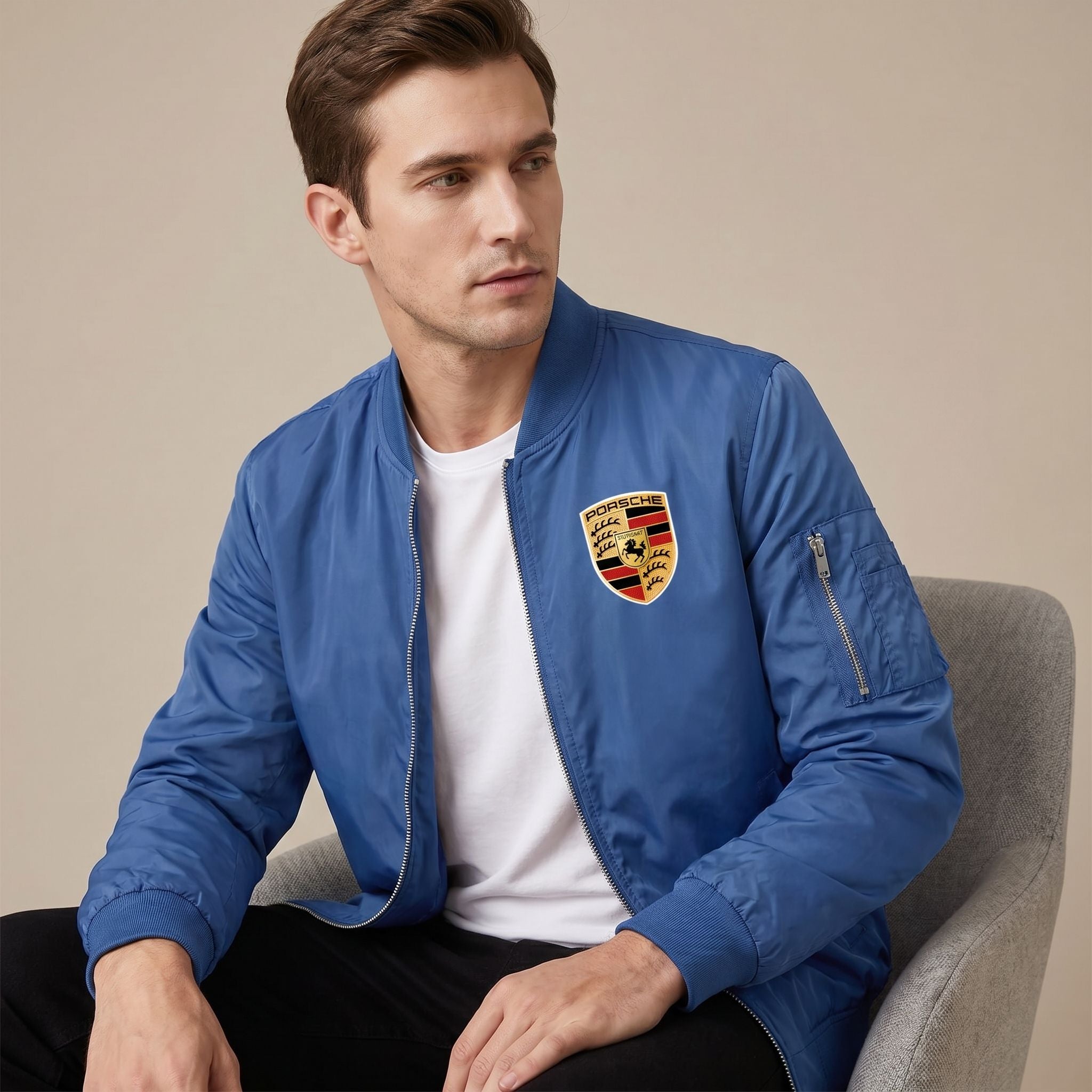 Indoor studio image of male model seated in a white Porsche bomber jacket, highlighting its textured fabric, structured fit, and versatile style.