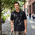 Male model wearing black Mercedes-Benz polo on city sidewalk with urban greenery and textured stone paving.