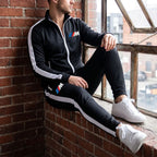 Casual scene of male model relaxing against brick wall in BMW M Dri-Fit Track Suit, highlighting relaxed fit and fabric details in natural light.