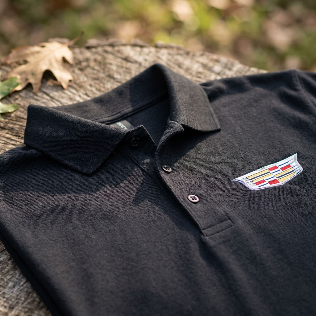 Close-up of men's Cadillac Car Dry Blend Polo showing textured cotton-polyester fabric, structured collar, and dyed-to-match buttons in natural outdoor setting.