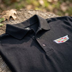 Close-up of men's Cadillac Car Dry Blend Polo showing textured cotton-polyester fabric, structured collar, and dyed-to-match buttons in natural outdoor setting.