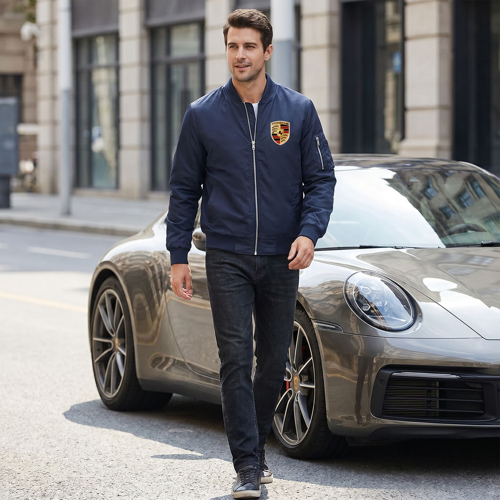 Full-body shot of male model wearing Porsche bomber jacket in city street with sleek car, emphasizing fit and stylish details during daylight.