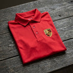 Studio shot of men's Porsche polyester polo laid flat on wooden surface, emphasizing smooth texture and vibrant color with minimal background.