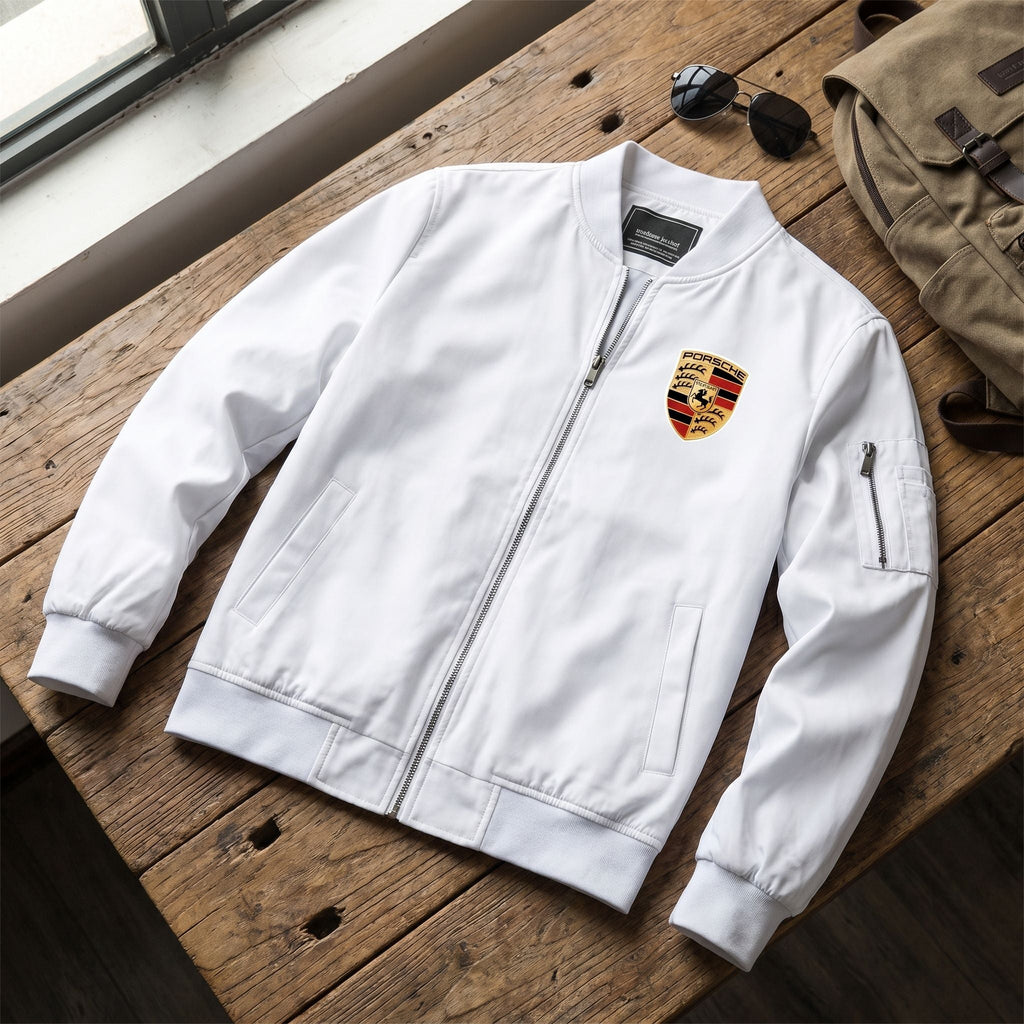 Flat-lay of men's Porsche bomber jacket with sunglasses and backpack on wooden surface.